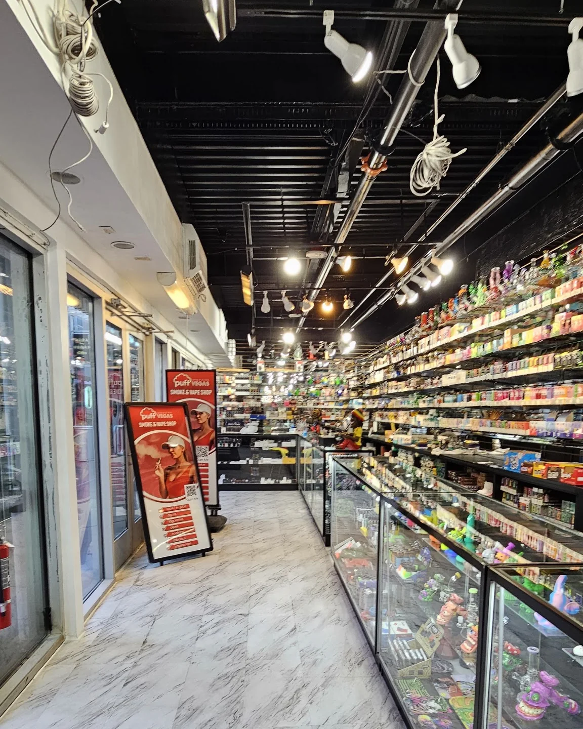 Looking up the aisle inside Puff Vegas: two Puff Vegas signs by the entrance, glass cases of pipes on the right, the wall of vapes behind them.
